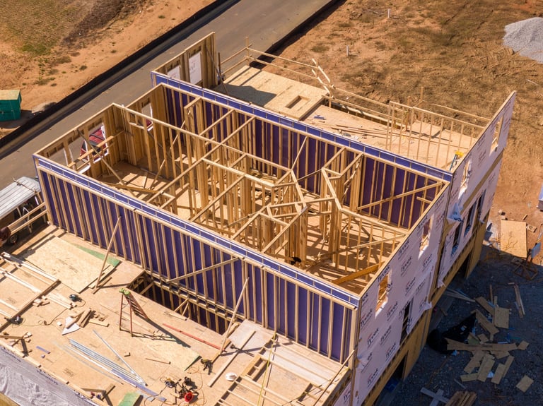 This high-angle shot captures a detailed look at the structural skeleton of a modern multi-story home. The image highlights the precise craftsmanship of the timber framing, including interior wall partitions and the subflooring of the upper levels.