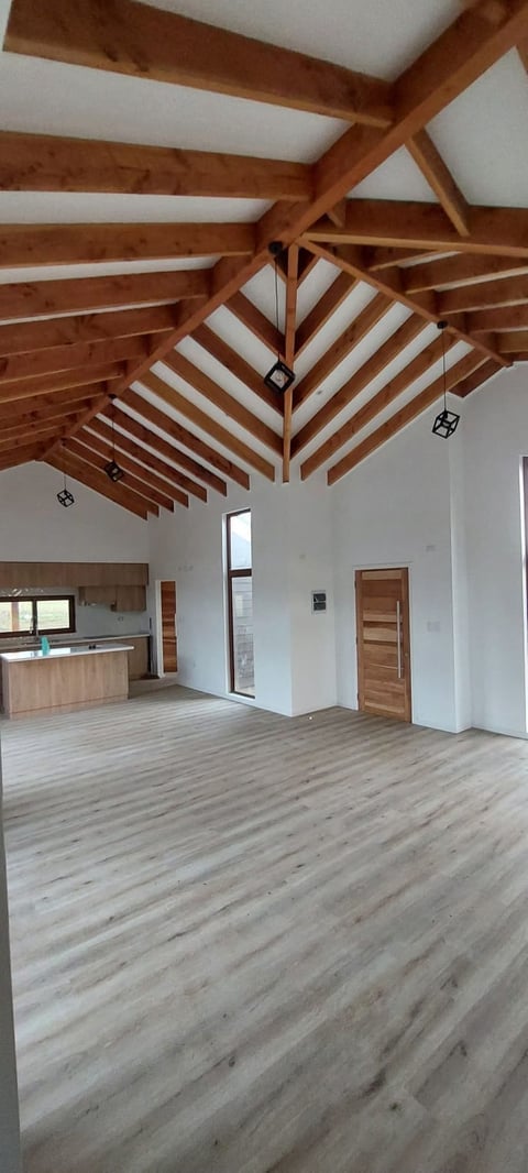 Interior of a modern open-plan space with exposed wooden beam ceiling, light wood flooring, white walls, kitchen area, and wooden doors