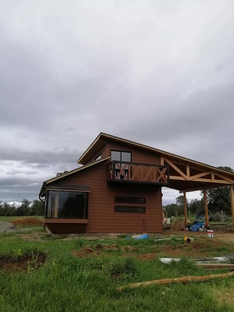 Brown wooden cabin with covered porch under construction in rural field with overcast sky