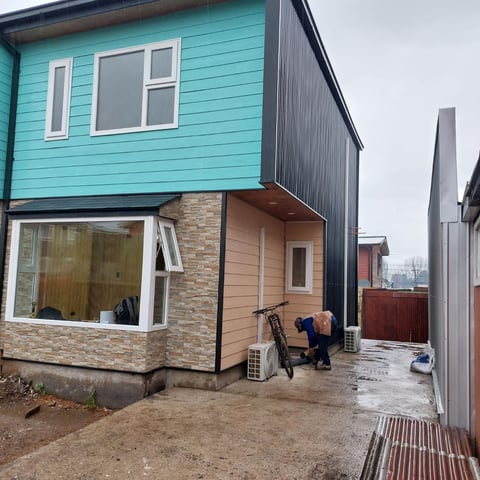 Modern two-story house with turquoise upper level and brick lower level, bicycle parked under covered entrance, paved patio area