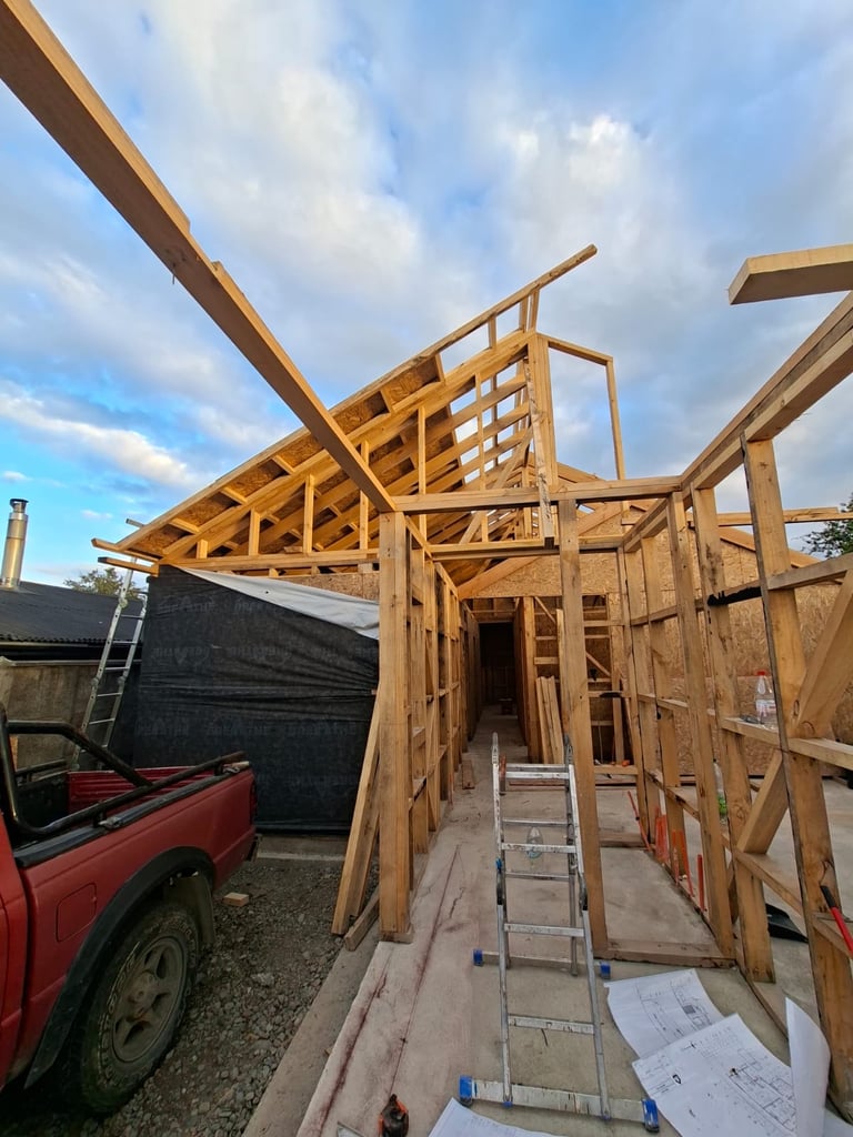 Wooden house frame under construction with exposed beams and trusses against a blue sky, with a red truck visible on the left