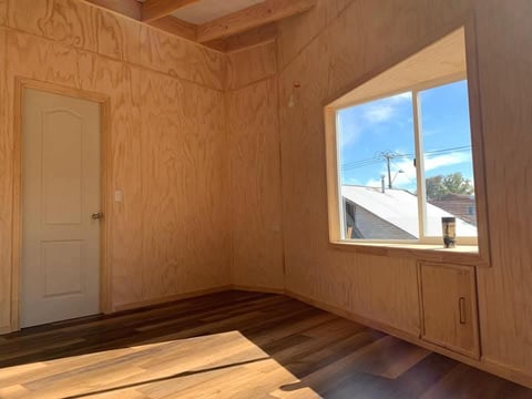 Empty room with plywood walls, wooden door, window showing outdoor view, and wooden flooring